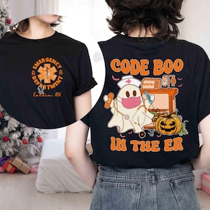 Könnte beinhalten: Schwarzes T-Shirt mit orangefarbenem und weißem Text und Grafiken. Die Vorderseite zeigt ein Notaufnahme-Logo und den Text "Emergency Department" und "Katie, RN". Auf der Rückseite befindet sich ein Geister-Krankenpfleger mit dem Text "Code Boo In The ER".