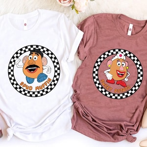 Può includere: Due magliette, una bianca e una malva, ciascuna con un personaggio di cartoni animati a forma di patata all'interno di un cerchio a scacchi bianco e nero. La maglietta bianca ha il testo "Spud Muffin", la maglietta malva "Sweet Potato".