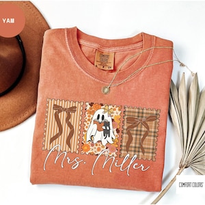 Op de afbeelding: Een koraalkleurig t-shirt met een afbeelding van een spook dat een boek leest met een kat en twee strikdesigns. De tekst "Mrs. Miller" is in cursieve letters onder de afbeelding geschreven. Een gouden ketting is over het shirt gedrapeerd.
