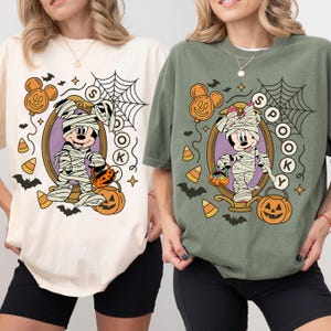 Op de afbeelding: Twee T-shirts, één crème en één olijfgroen, met een Halloween-ontwerp. Het ontwerp toont een in mummie gewikkelde Mickey Mouse met een pompoen, omringd door vleermuizen, snoep en het woord "Spooky".