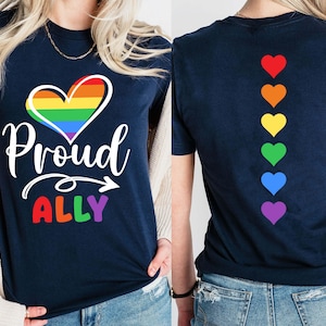Op de afbeelding: Marineblauw t-shirt met een regenbooghart en de woorden "Proud ALLY" in wit en regenboogkleuren. De achterkant van het shirt heeft een verticale lijn van regenboogkleurige harten. Een inclusief ontwerp.