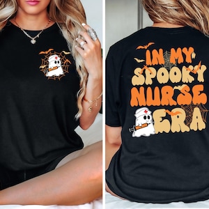 May include: Black t-shirt with a Halloween-themed design. The front features a ghost wearing a nurse's cap and holding a syringe, surrounded by a spider web. The back of the shirt says "IN MY SPOOKY NURSE ERA" in orange and tan lettering, with bats and a ghost.