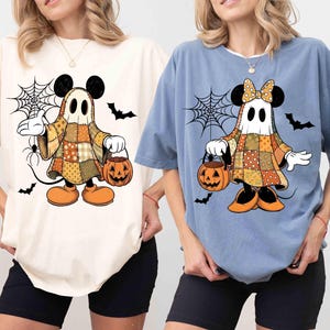 May include: Ivory and blue jean colored t-shirts featuring a Halloween-themed Mickey Mouse ghost design. The cartoon character is dressed in a patchwork costume and holding a jack-o'-lantern. Black bats and a spiderweb complete the spooky design.
