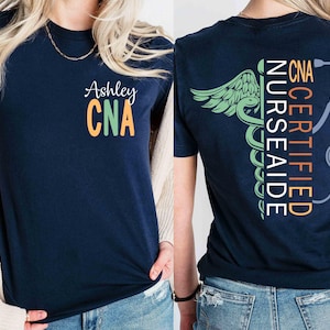 May include: Navy blue t-shirt with the name "Ashley" and "CNA" in orange and green on the front. The back of the shirt features a medical caduceus design with the words "CNA CERTIFIED NURSE AIDE" and a stethoscope.