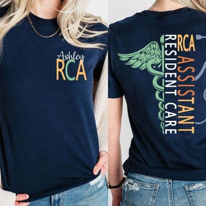Op de afbeelding: Marineblauw T-shirt met de tekst "Ashley RCA" op de voorkant. De achterkant van het shirt heeft de woorden "RCA ASSISTANT RESIDENT CARE" verticaal, samen met een medisch symbool en een stethoscoopafbeelding. Het shirt heeft een ronde hals.