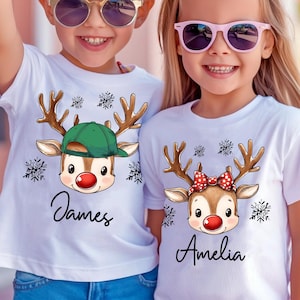 May include: Two children wearing white t-shirts with reindeer graphics. One reindeer wears a green cap, the other a red bow. The shirts have the names "James" and "Amelia" in script, with snowflake accents.