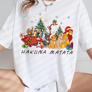 Puede incluir: Camiseta blanca con un diseño festivo de personajes de El Rey León. El gráfico incluye a Simba, Timón, Pumba y otros, con árboles de Navidad, copos de nieve y la frase "HAKUNA MATATA" en letras negras.