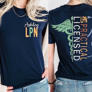 May include: Navy blue t-shirt with the name "Ashley" and "LPN" in green and orange on the front. The back of the shirt features a medical symbol, the words "PRACTICAL LICENSED LPN", and a stethoscope graphic.
