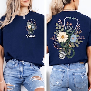 May include: Navy blue t-shirt featuring a floral design with a stethoscope and the word "Name". The design includes daisies, blue flowers, and green leaves. The back of the shirt has a similar design with a heartbeat line.