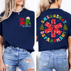 May include: Navy blue t-shirt with a festive ER design on the front, featuring a nurse's hat and the name "William." The back displays "EMERGENCY DEPARTMENT" around a red medical symbol with Christmas tree accents.