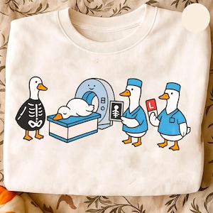 Könnte beinhalten: Weißes Sweatshirt mit einer Cartoon-Illustration von Enten in einer medizinischen Umgebung. Eine Ente ist ein Skelett, eine andere befindet sich in einem MRT-Gerät, und zwei Enten tragen OP-Kleidung und halten medizinische Bilder. Das Design hat blaue, schwarze und gelbe Akzente.