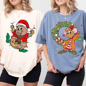 Puede incluir: Dos camisetas, una marfil y otra azul, con diseños de ratones de dibujos animados navideños. La camiseta marfil tiene un ratón con un gorro de Papá Noel y sujetando luces de cadena. La camiseta azul tiene un ratón con una corona y un bastón de caramelo.