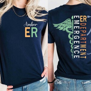 Pode incluir: Camiseta azul marinho com "Amber" e "ER" em verde e laranja na frente. A parte de tr&aacute;s apresenta um design de caduceu m&eacute;dico e estetosc&oacute;pio com as palavras "ER DEPARTMENT EMERGENCY" em texto vertical. Um artigo de vestu&aacute;rio com tema de sa&uacute;de.