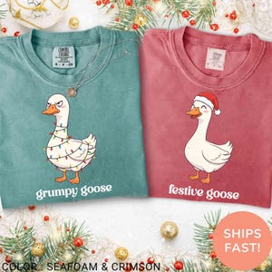 May include: Two Comfort Colors t-shirts, one seafoam green and one crimson. The seafoam shirt features a grumpy goose with Christmas lights, the crimson shirt has a festive goose wearing a Santa hat. Both shirts have text below the goose.