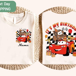 Op de afbeelding: Crèmekleurig T-shirt met een kleurrijke afbeelding van de Cars-personages Bliksem McQueen en Takel, met de tekst "It's My Birthday". Het ontwerp bevat verjaardagselementen zoals een taart, ballonnen en geblokte vlaggen. Het shirt heeft een ontwerp aan de voor- en achterkant.