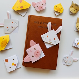 Può includere: Una collezione di segnalibri colorati a tema animale con un maiale, un coniglio, un pulcino e un cavallo. I segnalibri sono piegati a forma di angolo e sono posizionati su un libro marrone intitolato "Alice nel Paese delle Meraviglie" di Lewis Carroll.