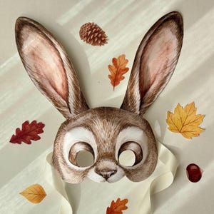 May include: A watercolor-style bunny mask with long ears, brown fur, and white eye openings. The mask is surrounded by autumn leaves, a pine cone, and a ribbon. The mask is suitable for a costume or party.