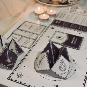 May include: A black and white witch spell card game with a fortune teller style paper fortune. The fortune teller is folded into a pyramid shape with the words "Broom", "Potion", and "Wand" visible. The card game has a black background with white stars and the words "Witch Spell Cards".