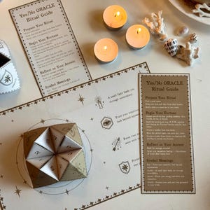 Puede incluir: Un juego de ritual de oráculo beige con un adivino, tarjetas de guía y velas encendidas. El adivino es una manualidad de papel con números. Las tarjetas de guía tienen el texto "Yes/No ORACLE Ritual Guide".