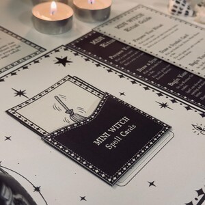 May include: A black and white card game called "Mini Witch Spell Cards". The cards are in a black pocket with a white border and a white star pattern. The card in the pocket shows a black and white illustration of a broom.