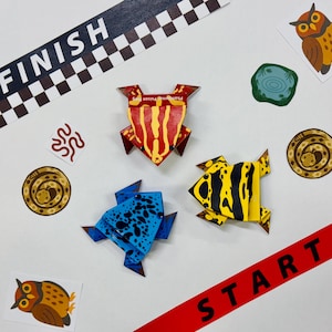 Puede incluir: Tres ranas de origami en rojo, azul y amarillo con rayas negras están dispuestas sobre una superficie blanca. La palabra "START" está en una cinta roja y "FINISH" en una cinta a cuadros. También hay pegatinas de un búho, una serpiente y un estanque.