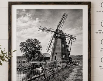 Op de afbeelding: Een zwart-wit wandkunstprint met een rustieke windmolen bij een waterkant en een houten hek. De scène bevat een grote boom en een klein gebouw op de achtergrond onder een bewolkte lucht, ingelijst in een donkere houten lijst.