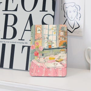 May include: A rectangular coaster featuring a cozy interior scene with a table set for tea. The table is covered with a pink tablecloth and adorned with a teacup, teapot, and open book. The background shows a window with curtains and a bookshelf.