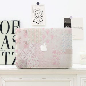 Op de afbeelding: Een roze en witte patchwork laptophoes met een bloemen- en strikpatroon. De hoes heeft het Apple-logo en staat op een wit oppervlak. Decoratieve kunst en een kaars staan op de achtergrond.