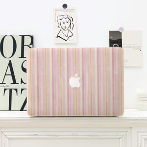 May include: A pink and white striped laptop case with the Apple logo. The case features vertical stripes in pink, white, and gold. The laptop case is on a white shelf, with decorative art and a candle in the background.