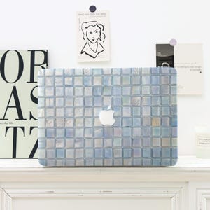 May include: A laptop featuring a mosaic tile pattern in blue, white, and grey. The Apple logo is in the centre. The laptop is on a white surface, with decorative items in the background.