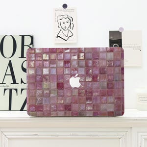 Op de afbeelding: Een laptophoes met een raster van vierkante, iriserende tegels in verschillende tinten paars, magenta en mauve. Het oppervlak heeft een glanzende, reflecterende afwerking met een centrale uitsparing die het zilveren Apple-logo onthult.