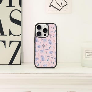 May include: A black iPhone case with a pink background featuring a repeating pattern of blue cats, leaves, and floral elements. The case has a black border and a camera cutout.