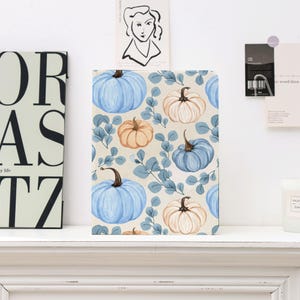 Può includere: Un taccuino con un design a tema autunnale con zucche ad acquerello nei toni del blu, arancione e crema, intervallate da foglie verde-blu, su uno sfondo crema. Il taccuino è esposto su una mensola bianca.
