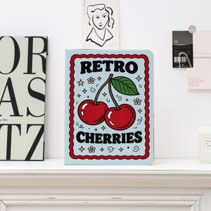 May include: A light blue notebook with a red border and the words "RETRO CHERRIES" in a retro font. The cover features a pair of red cherries with a green leaf. Decorative stars and flowers surround the text and cherries.