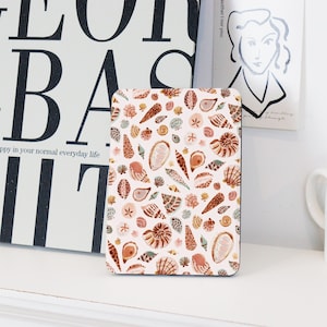 May include: A square coaster with a white background and a pattern of brown, pink, and blue seashells.