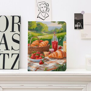 Może przedstawiać: Notatnik z ilustracją sceny piknikowej. Obraz przedstawia kosz z rogalikami, miskę truskawek i napoje na kraciastej kołdrze. Tło przedstawia zielony krajobraz i błękitne niebo.