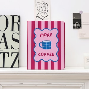 May include: A pink and red striped notebook with the words "MORE COFFEE" and a blue coffee cup graphic on the front. The notebook is displayed on a white shelf with other decorative items.