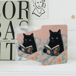 Könnte beinhalten: Eine transparente Handyhülle und eine Telefonkarte mit einer schwarzen Katze, die ein Buch liest. Die Katze sitzt auf einem Kissen mit Blumenmuster. Der Hintergrund ist hellrosa.