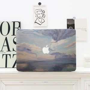 Op de afbeelding: Een laptop met een hoes met een zeelandschapontwerp in blauwe, grijze en beige tinten. Het Apple-logo is zichtbaar. De laptop staat op een wit oppervlak.