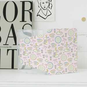 May include: A clear phone case with a pink patterned insert. The pattern includes teacups, tiered cake stands, and bows in pastel colours. The case is designed to fit a phone. The design has a whimsical aesthetic.