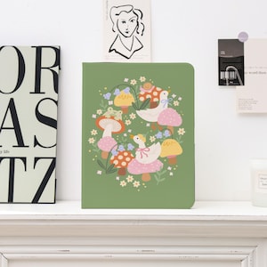 Op de afbeelding: Een groen notitieboek met een grillige illustratie van ganzen, paddenstoelen en een kikker. Het ontwerp toont witte ganzen met roze strikken, rood-wit gestippelde paddenstoelen en een kikker op een paddenstoelenhoed. Het notitieboek ligt op een witte plank.