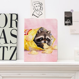 May include: A pink notebook with a watercolour illustration of a raccoon in a yellow crisp packet. The raccoon is eating a crisp. The notebook is on a white shelf, with other decorative items in the background.