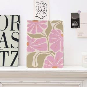 May include: A rectangular notebook with a floral design. The cover features large pink flowers with orange centres and olive green leaves on a cream background. The notebook is on a white shelf.