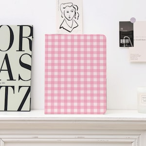 May include: A pink and white gingham pattern notebook. The notebook is rectangular and stands upright on a white shelf. Other decorative items are visible in the background, including a black and white art print.