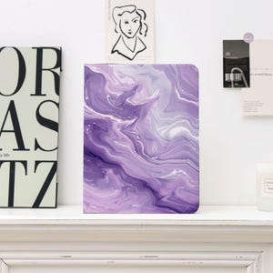Può includere: Un taccuino rettangolare con un design marmorizzato viola e bianco. La copertina presenta motivi a vortice di tonalità viola chiare e scure, creando un'estetica astratta fluida. Il taccuino è esposto su una mensola bianca.