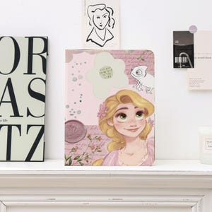 May include: A pink journal featuring a cartoon illustration of a princess with long blonde hair and green eyes. The journal has a floral design, a white flower-shaped sticker with text, and a drawing of a chameleon.