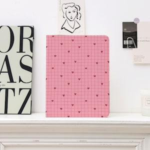May include: A pink notebook with a grid pattern and small red hearts. The notebook is rectangular and stands upright on a white shelf. Other decorative items are visible in the background.