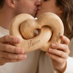 Puede incluir: Escultura de madera en forma de corazón con dos corazones entrelazados. La escultura es de color marrón claro y tiene los nombres "Lucien" y "Evelyn" grabados. Un pareja se besa en el fondo.