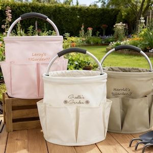 K&ouml;nnte beinhalten: Drei Gartenwerkzeug-Organizer aus Canvas in Rosa, Creme und Beige, jeweils mit Metallgriff und mehreren Au&szlig;entaschen. Auf der rosa Tasche steht "Leroy's Garden". Auf der cremefarbenen Tasche steht "Giselle's Garden". Auf der beigefarbenen Tasche steht "Harold's Garden".