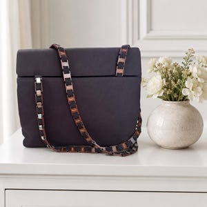 May include: A dark gray handbag with a rectangular shape and a flap closure. The bag features a chain strap with brown and silver accents. A small vase with white flowers sits to the right of the bag.